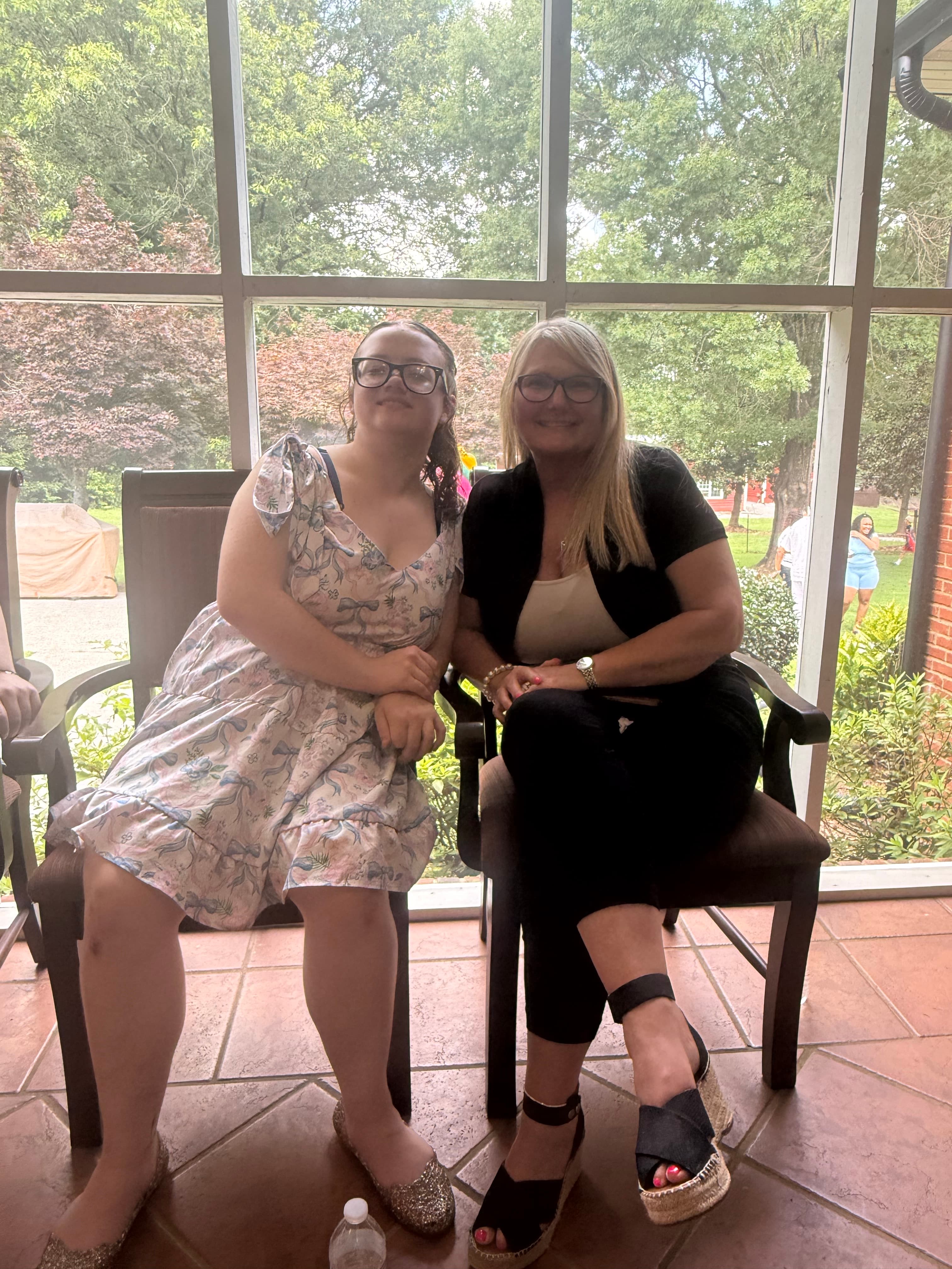 Two women wearing glasses sit in chairs on a screened porch with green trees behind.