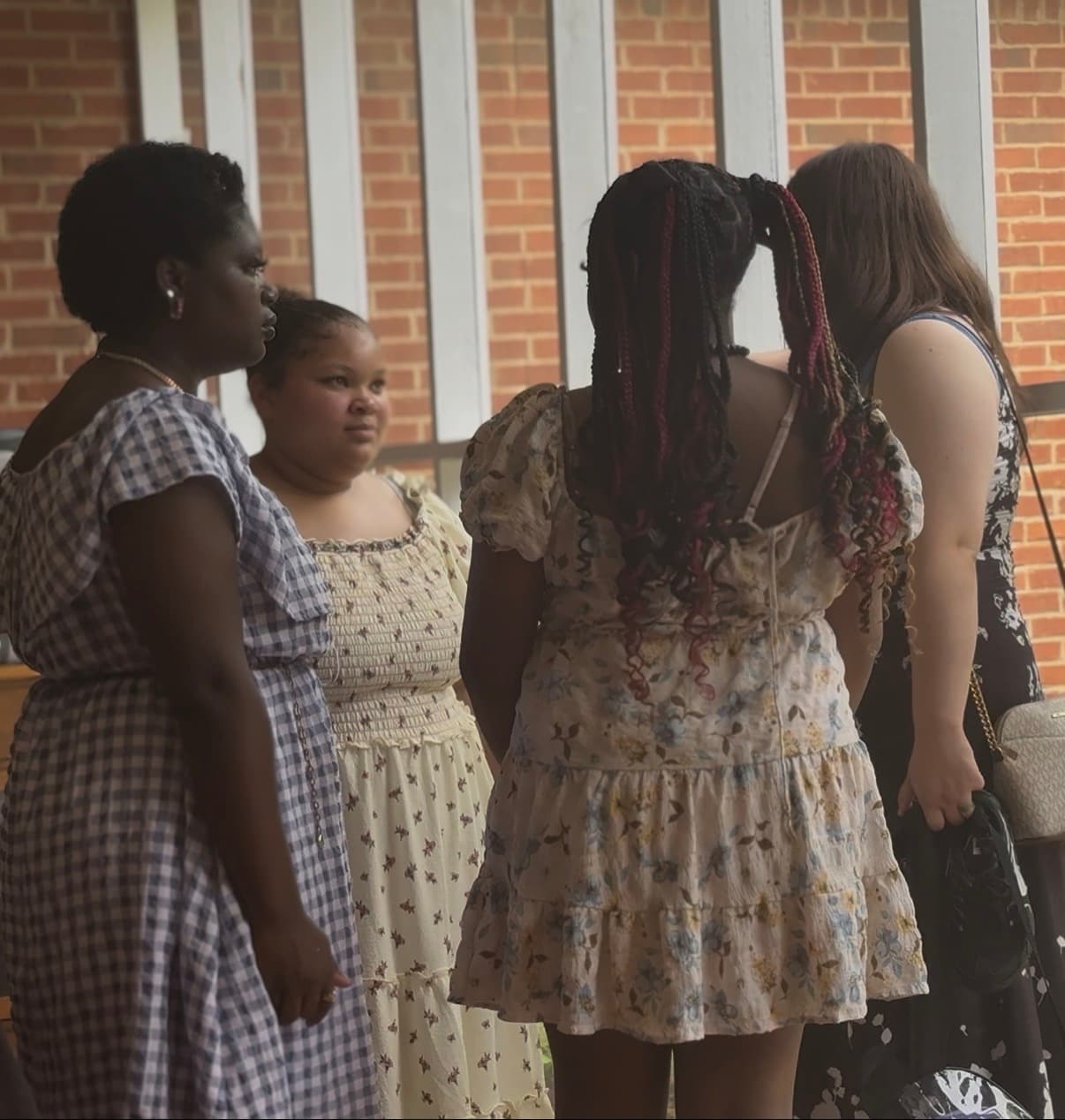 Four women in patterned summer dresses stand together outdoors against a brick and pillar background.
