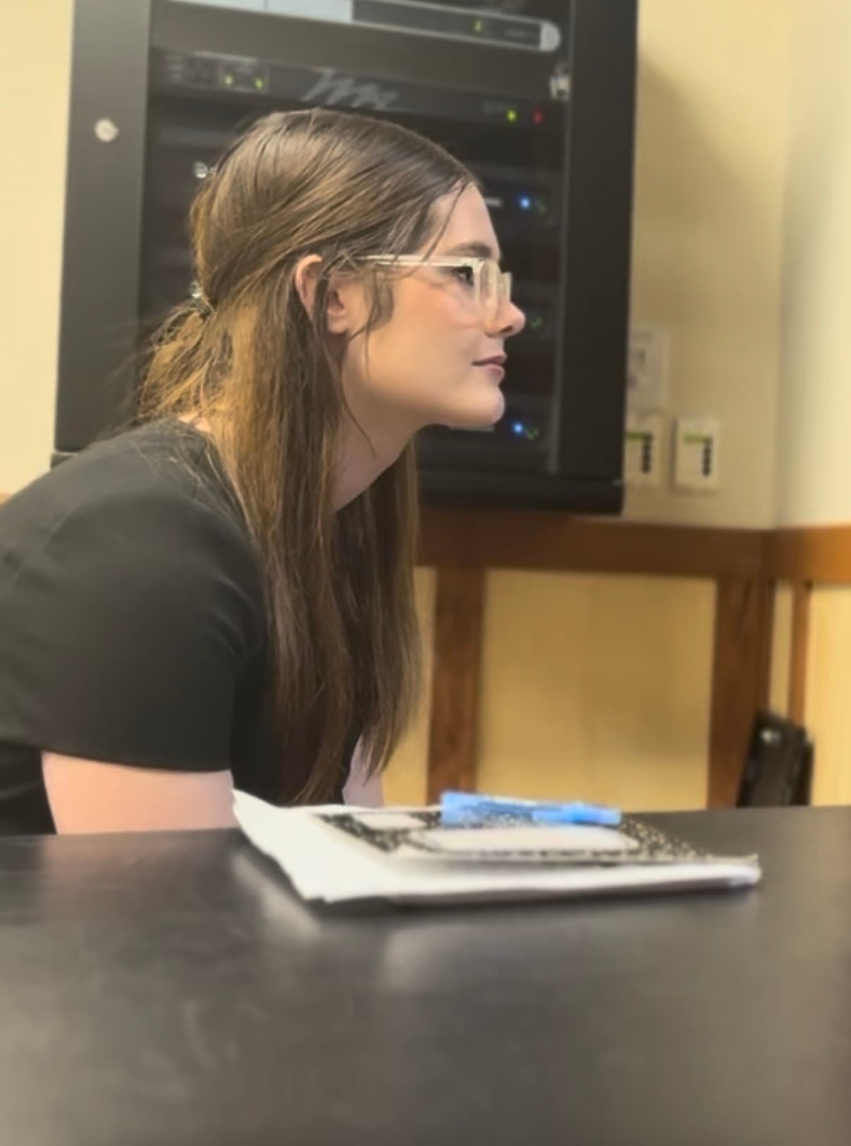 Woman with long brown hair and glasses sits at a desk before a server rack.