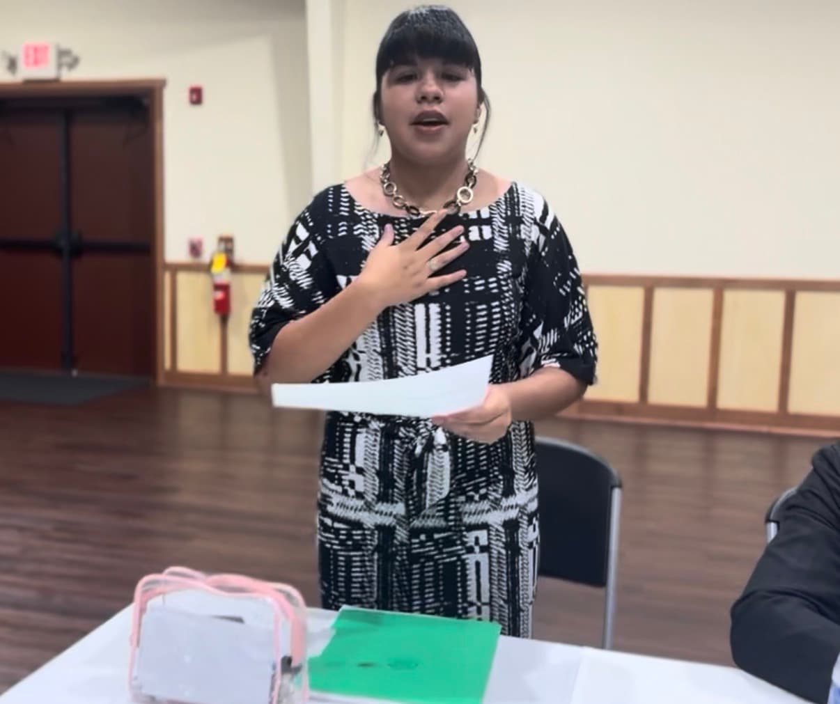 Woman in patterned dress speaks, holding a paper with one hand over her heart.