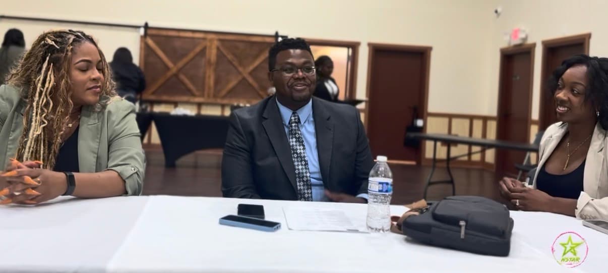 Three smiling professionals in business attire sit at a white table during a meeting.