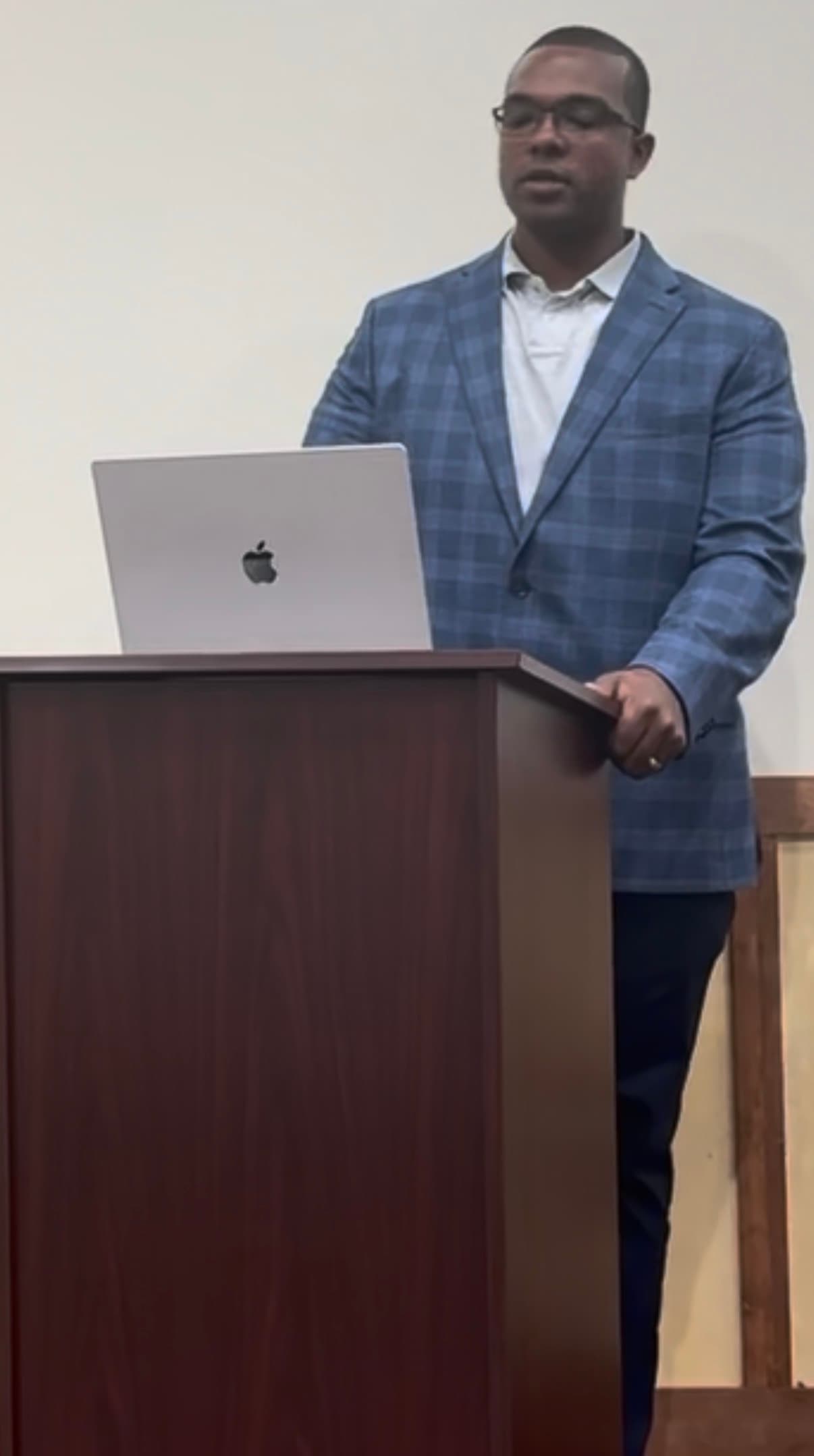 A Black man in a blue plaid blazer stands behind a podium with a laptop.