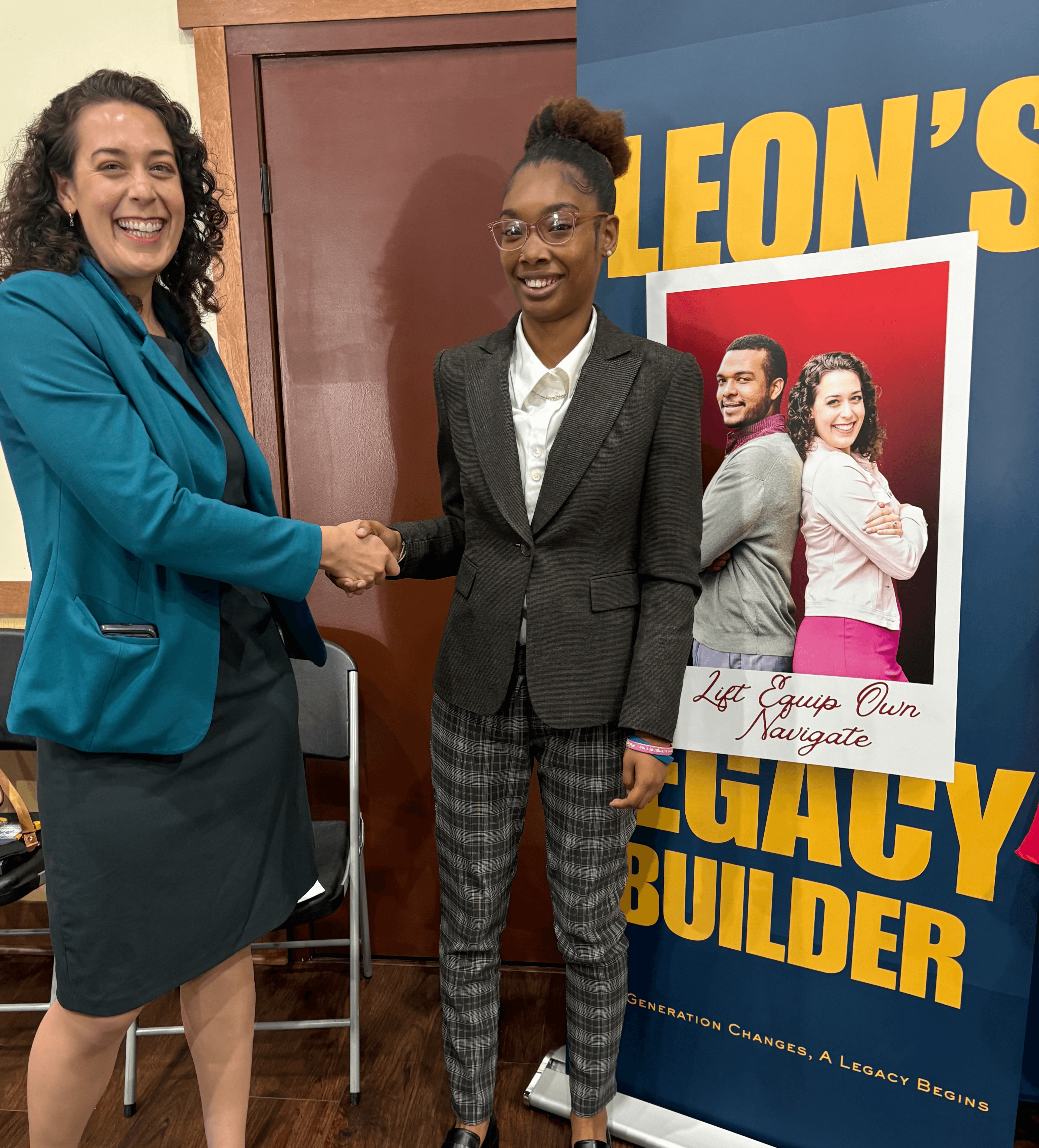 Two smiling women in business attire shake hands beside a Leon's Legacy Builder banner.